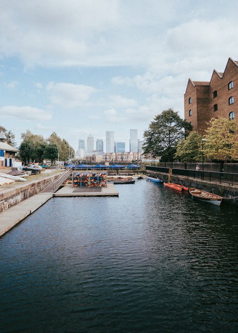 A wide view of a narrow waterway in Canary Wharf, showing a concrete walkway on the left with stacked cardboard boxes and orange pallets nearby, and several small wooden boats moored along the right side; modern high-rise skyscrapers are visible in the background under a partly cloudy sky, with trees lining the walkway and a metal railing separating the water from the urban environment. The scene captures elements involved in house removals or furniture transport in a high-rise flats area, with the setting indicating a relocation process possibly involving loading or unloading activities, supported by the professional removal service offered by Man with Van Docklands.
