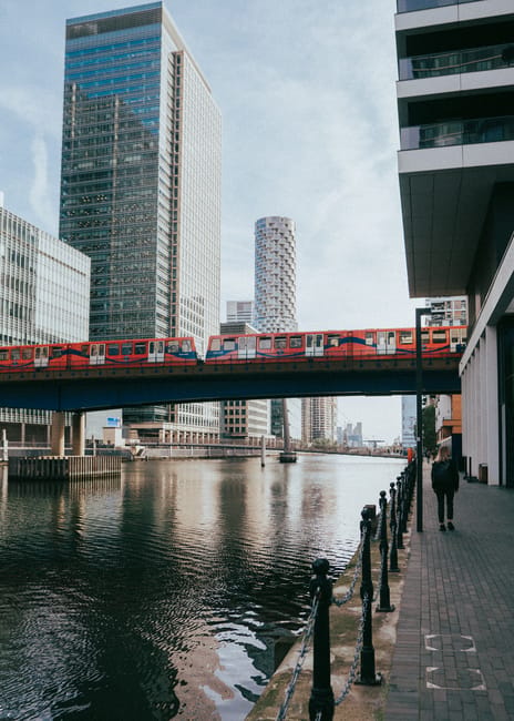The image shows a section of a high-rise residential building on the right side with an overhanging balcony. Below is a paved walkway separated from a canal by black metal railings. A person dressed in dark clothing is walking along the pathway. In the background, a red train is passing over a bridge spanning the canal, supported by concrete pillars. Surrounding the waterway are tall, modern office and residential buildings with glass facades, reflecting the sky. The scene is captured during daylight with clear weather, and the atmosphere suggests an urban environment suitable for house removals or furniture transport around the Canary Wharf area, as detailed in the Man with Van Docklands removals guide.