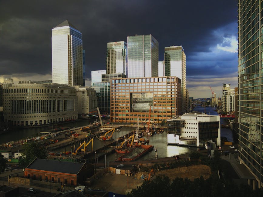 A wide view of a narrow waterway in Canary Wharf, showing a concrete walkway on the left with stacked cardboard boxes and orange pallets nearby, and several small wooden boats moored along the right side; modern high-rise skyscrapers are visible in the background under a partly cloudy sky, with trees lining the walkway and a metal railing separating the water from the urban environment. The scene captures elements involved in house removals or furniture transport in a high-rise flats area, with the setting indicating a relocation process possibly involving loading or unloading activities, supported by the professional removal service offered by Man with Van Docklands.