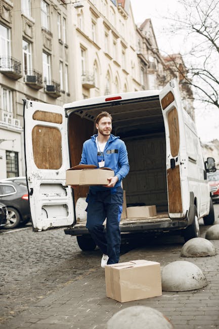 A man wearing a blue jacket and work trousers is standing outside a white box van with its rear doors open on a cobblestone street in an urban residential area during daylight hours. He is carrying a