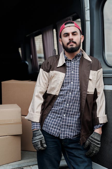 A man with a beard, wearing a red cap backwards, a beige and dark brown jacket over a blue checked shirt, and grey gloves, standing inside a van during a home relocation process. Behind him, there are stacked cardboard boxes of various sizes, some wrapped in plastic or packing tape, indicating packing for moving. The van's interior appears spacious, with a dark ceiling and side panels, and the scene is lit with natural daylight coming through the windows. This image captures a moment of furniture transport or packing during a professional move, reflecting the logistical process involved in home removals. The presence of the boxes and the loading environment aligns with the services offered by Man with Van Docklands, including urgent or same-day house removals in the Isle of Dogs area.