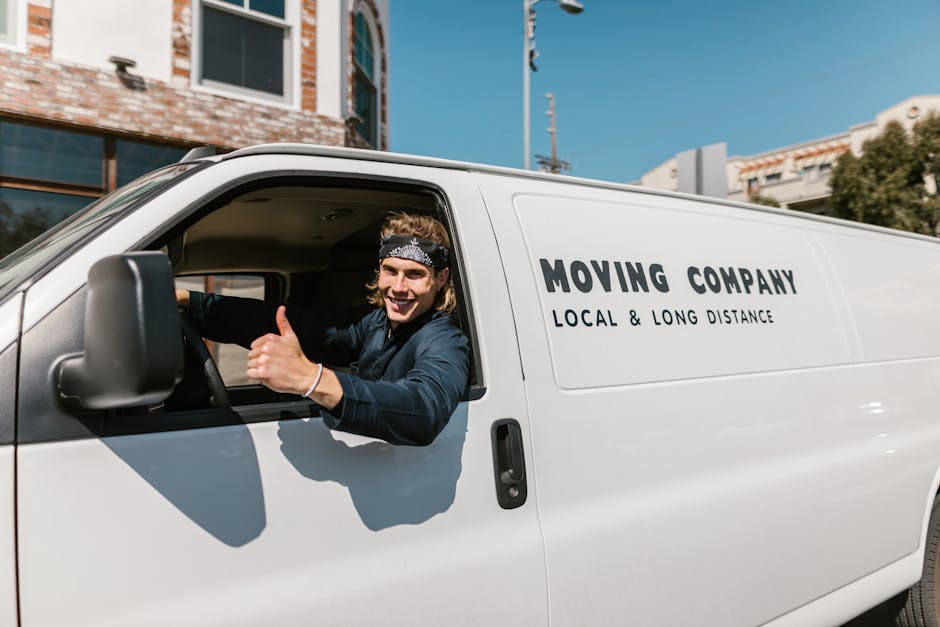 A smiling young man with long, wavy hair, wearing a black and white bandana and a dark jacket, is sitting in the driver's seat of a white moving van with the words 'MOVING COMPANY LOCAL & LONG DISTANCE' printed on the side. The vehicle is parked outdoors on a clear day, with modern buildings, trees, and a blue sky visible in the background. The man is giving a thumbs-up gesture through the open window, indicating readiness for a home relocation or furniture transport. Inside the vehicle, some interior details like the steering wheel and part of the dashboard are visible. The scene suggests an active moving process, with the van prepared for loading or unloading household items, with the driver appearing approachable and professional, supporting services like packing and loading for house removals, as conducted by Man with Van Docklands.