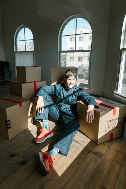 A man with a beard, wearing a red cap backwards, a beige and dark brown jacket over a blue checked shirt, and grey gloves, standing inside a van during a home relocation process. Behind him, there are stacked cardboard boxes of various sizes, some wrapped in plastic or packing tape, indicating packing for moving. The van's interior appears spacious, with a dark ceiling and side panels, and the scene is lit with natural daylight coming through the windows. This image captures a moment of furniture transport or packing during a professional move, reflecting the logistical process involved in home removals. The presence of the boxes and the loading environment aligns with the services offered by Man with Van Docklands, including urgent or same-day house removals in the Isle of Dogs area.