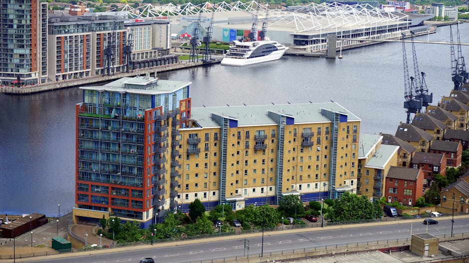 An aerial view of a residential building situated next to a river, with a parking area featuring several cars in front. The building has a modern design with multiple balconies, large windows, and a colorful façade combining beige, blue, and red panels. Behind the building, there are commercial structures, including a large stadium with a distinctive white roof supported by a network of steel beams, along with cranes and construction equipment on the waterfront. The river contains a docked white yacht, and the overall environment shows a mix of urban residential and commercial properties. The scene is well-lit with natural daylight, highlighting the details of the building's structure and the surrounding infrastructure, reflecting typical elements involved in home relocation or furniture transport services provided by Man with Van Docklands.