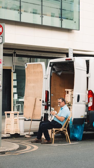 A man with glasses and a light blue shirt is seated on a wooden chair outside a white van, which is parked on a city street in front of a building with large windows and a glass balcony. The van has its rear doors open, revealing packed cardboard boxes and wrapped furniture inside, indicating an ongoing home relocation process. Several wooden pallets and a large wooden panel are propped against the building's exterior near the van. The street includes a curb and a no-parking sign. The scene appears to be part of a professional removals service, with the man resting during the loading or unloading phase, consistent with house removals or furniture transport activities carried out by Man with Van Docklands.